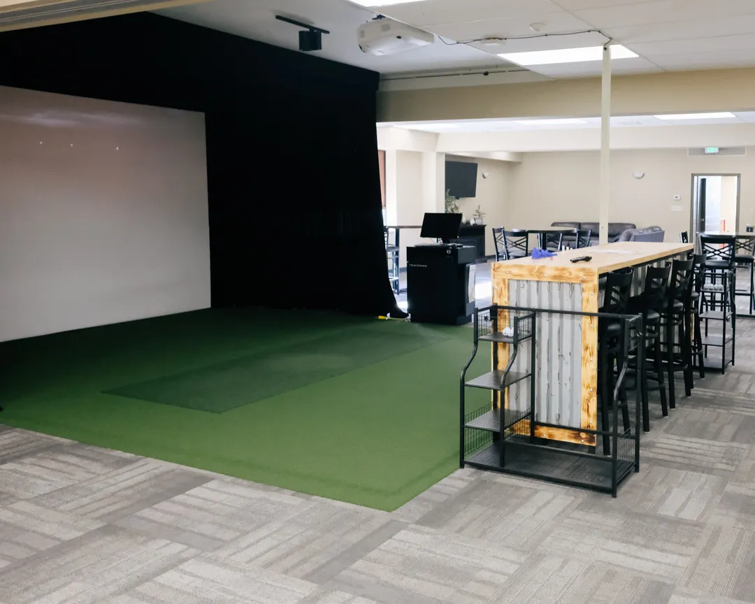 Golf simulator with a large white projector screen, computer equipment, projector, and a green play surface.