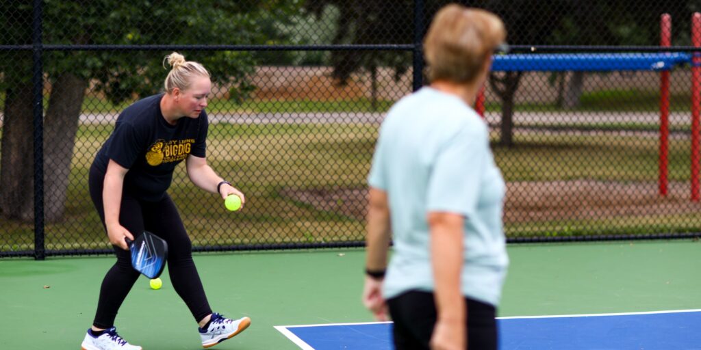 Woman with blond hair and a black t-shirt prepares to serve during a pickleball game on an outdoor court.