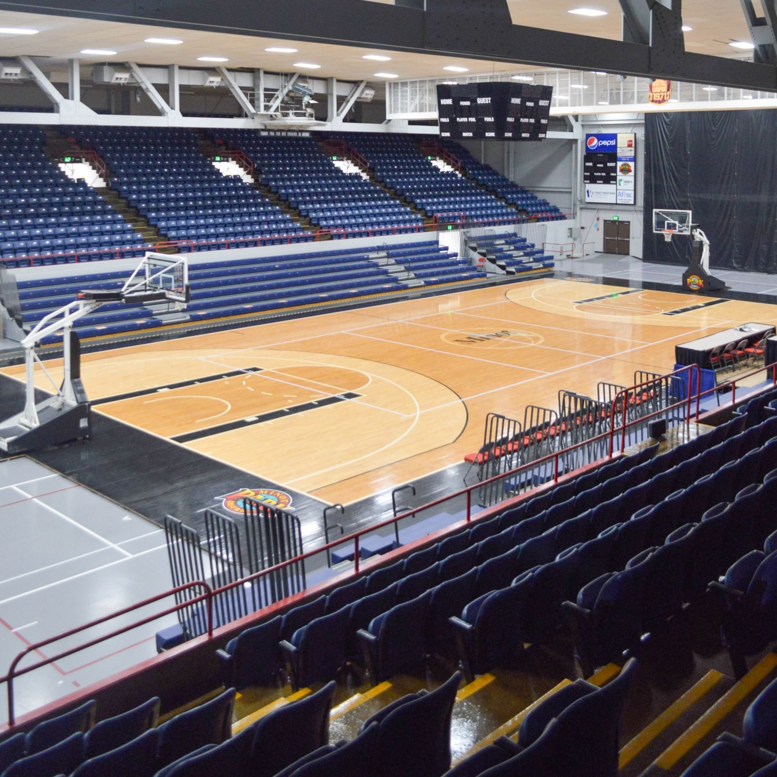 Interior view of a basketball court with blue seating, hoop, scoreboard, and overhead lighting.