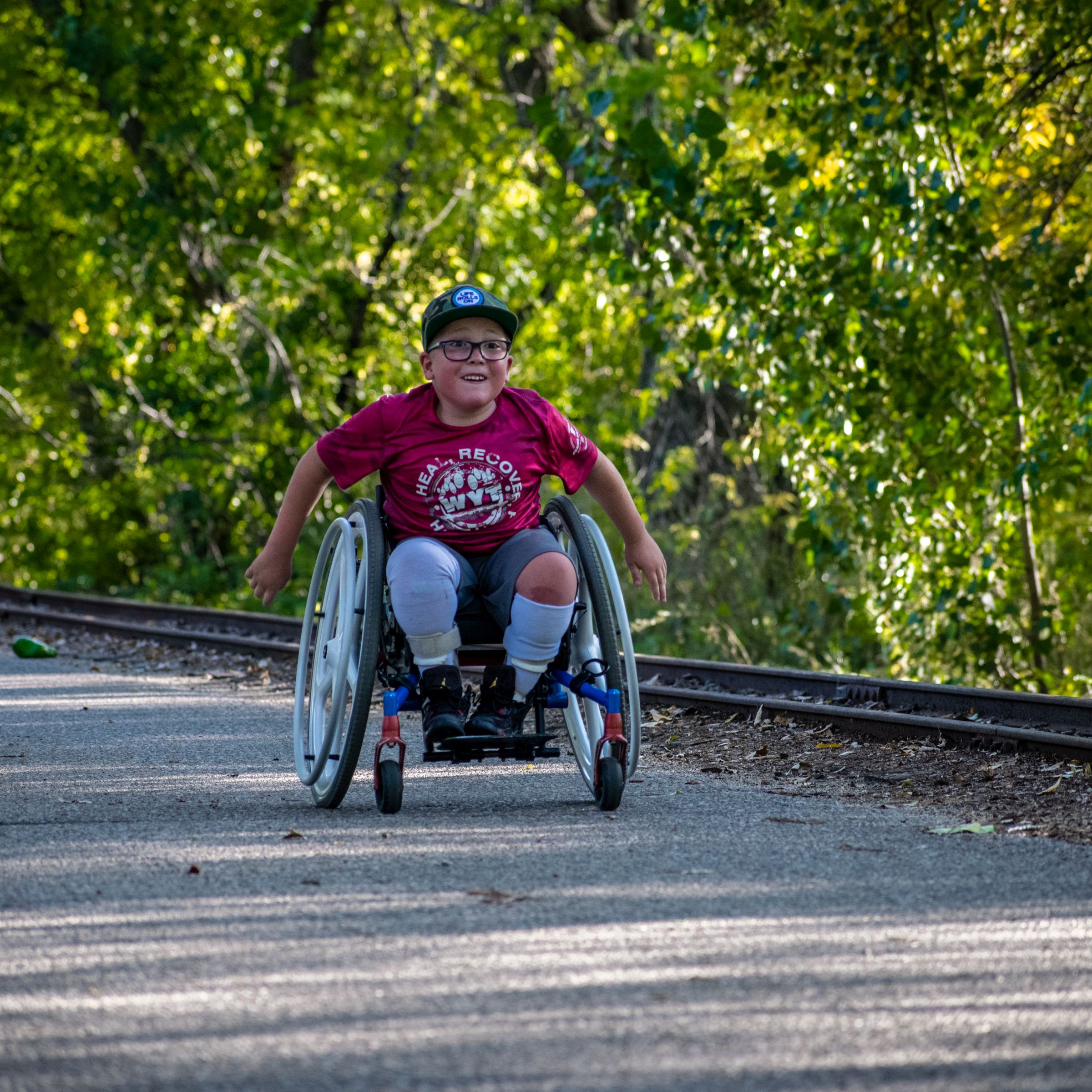 Young boy in a wheelchair smiles while moving down a paved path beside railroad tracks in bright daylight.