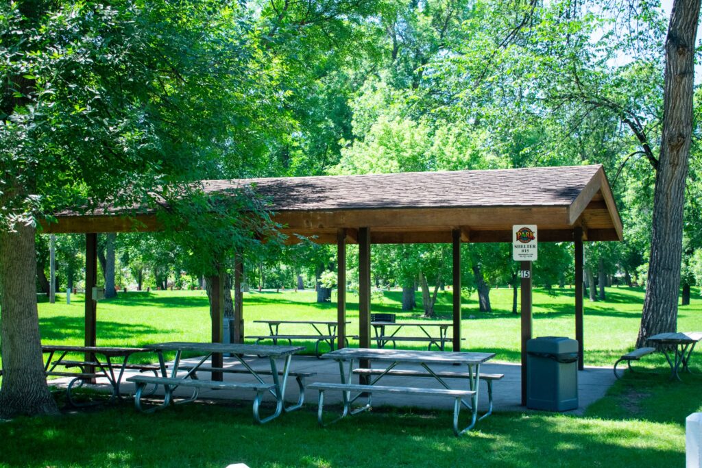 Oak Park shelter 15 with picnic tables under a covered structure, surrounded by green grass and trees, providing a shaded picnic spot.