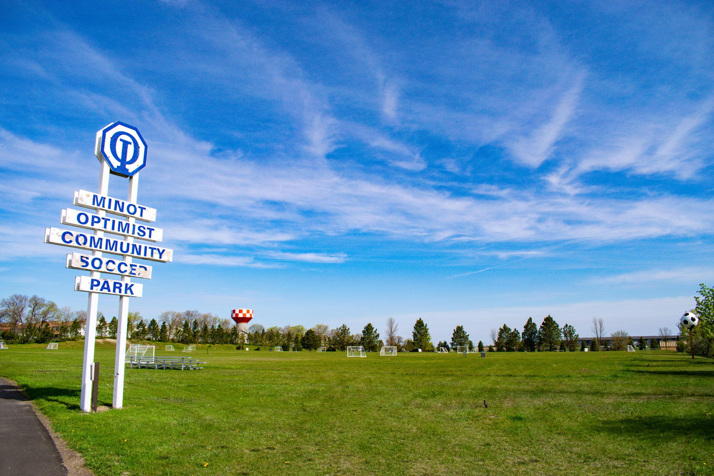 A large white and blue sign for the Minot Optimist Community Soccer Park, with several soccer fields in the background.