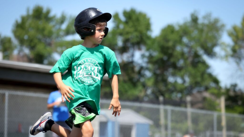 A young player wearing a batting helmet and a green Minot Park District t-shirt runs the bases during a game.