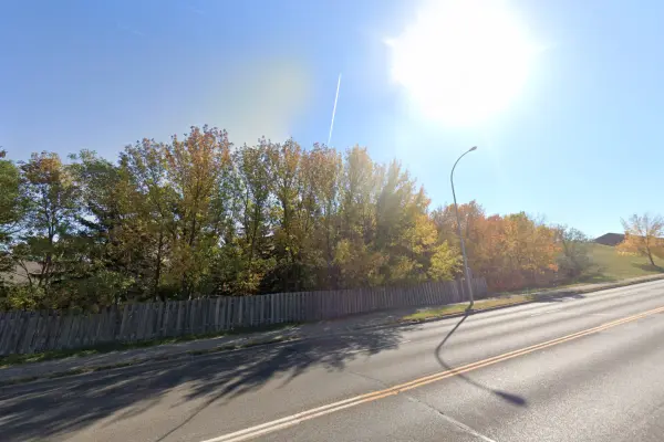 A sidewalk and street go up a hill with trees in the background showing red, yellow and green leaves of autumn under a cloudless sunny sky.
