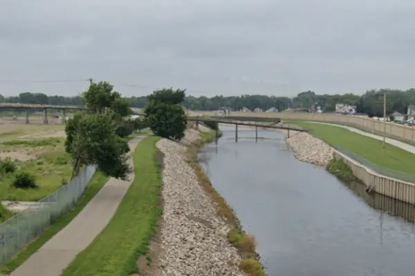 A narrow river flows between rocky banks with a paved walking path and green grass under an overcast sky.