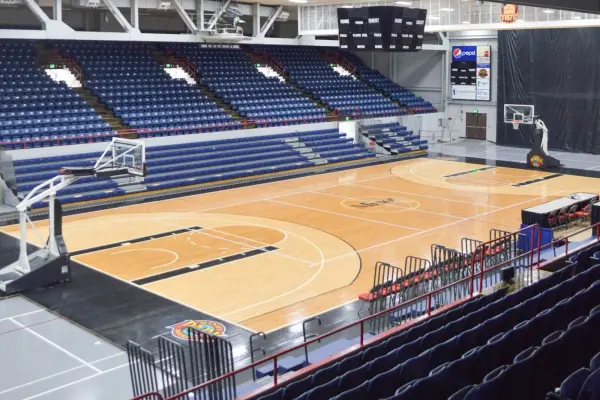Interior view of a basketball court with blue seating, hoop, scoreboard, and overhead lighting.