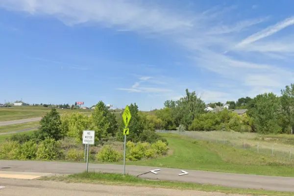 Google Maps screenshot of the BYP 83 – 4th Ave NW, a paved path that runs next to a street, with green space and trees in the background under a blue sky with wispy white clouds.