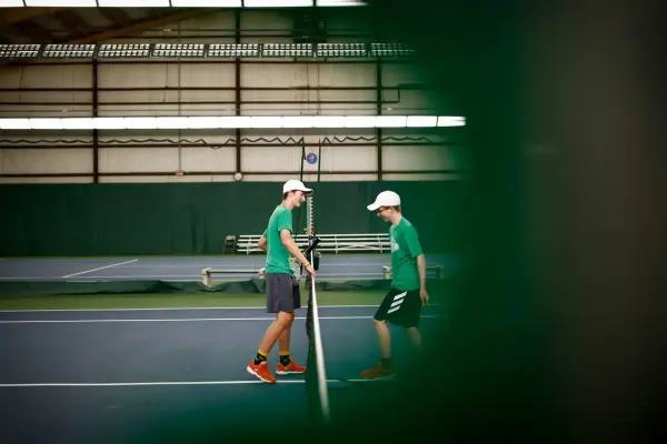 Two young men in green shirts and white caps walk past each other at the net on an indoor tennis court.