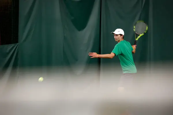Young man in green shirt and white cap hits a tennis ball with a racquet against a dark green backdrop.