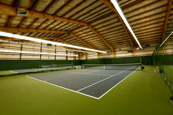 Indoor tennis court with grey surface, green surroundings, and bright overhead fluorescent lighting.