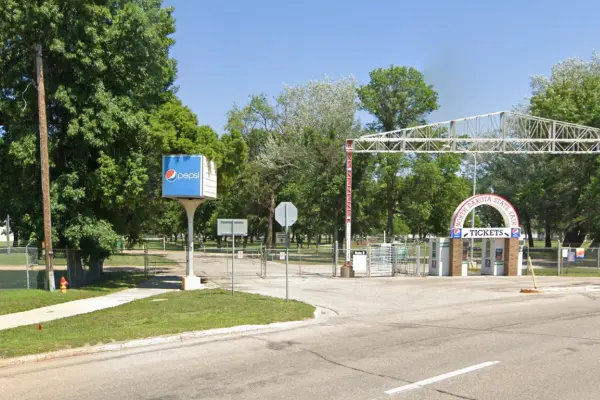 Gate to the North Dakota State Fairgrounds in Minot, ND. Signage to purchase tickets under an arched entrance, with trees and paved paths nearby.