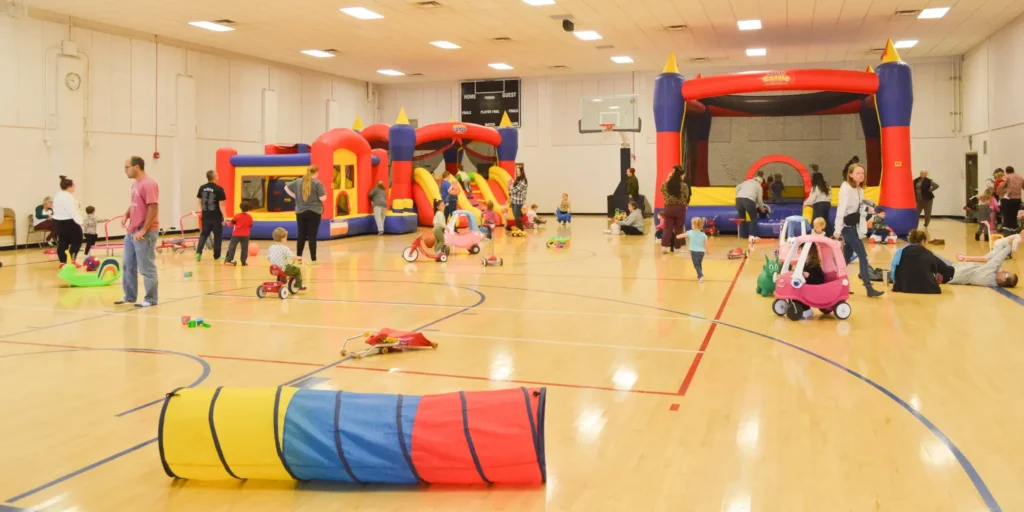 Children and adults playing with bounce houses and ride-on toys inside a brightly lit indoor gymnasium.