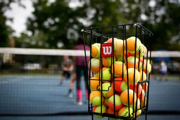A basket filled with yellow and orange tennis balls with a blurred image of a tennis court in the background.