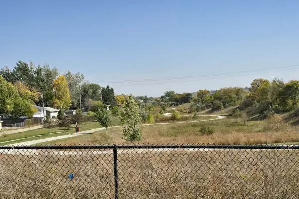Winding paved path leads through a grassy park with autumn trees under a clear blue sky, viewed behind a chain-link fence.