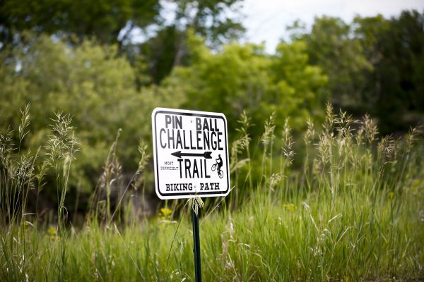 White metal trail sign reading Pin Ball Challenge Trail Biking Path standing in tall green grass.
