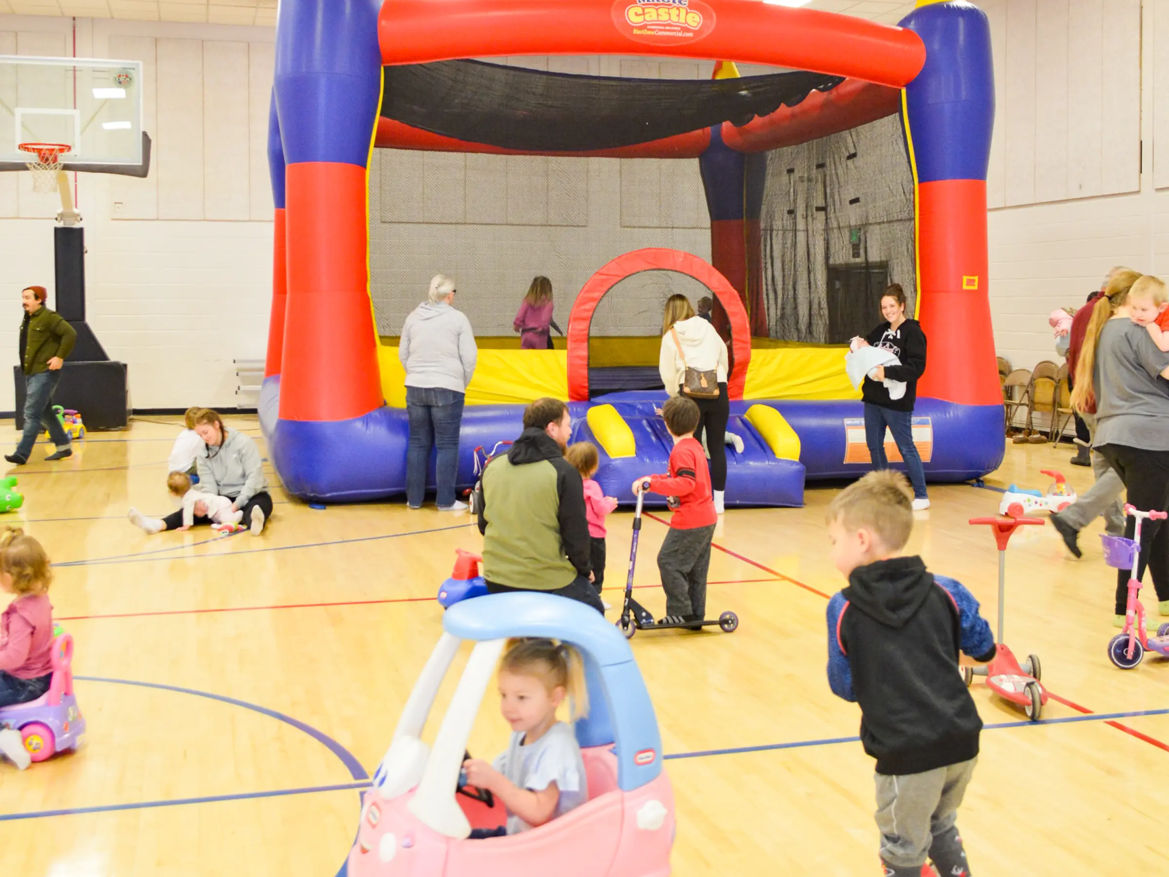 Children and adults playing with bounce houses and ride-on toys inside a brightly lit indoor gymnasium.