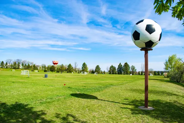 Large soccer ball statue stands on a grassy field with soccer goals under a blue, cloudy sky.