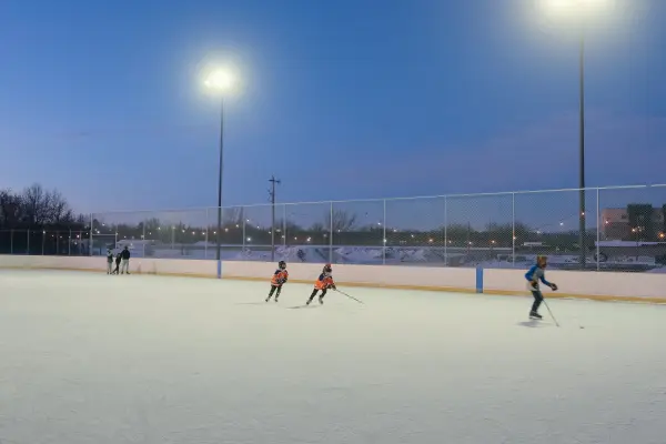 Children in hockey gear skating on an outdoor ice rink under bright stadium lights at twilight.