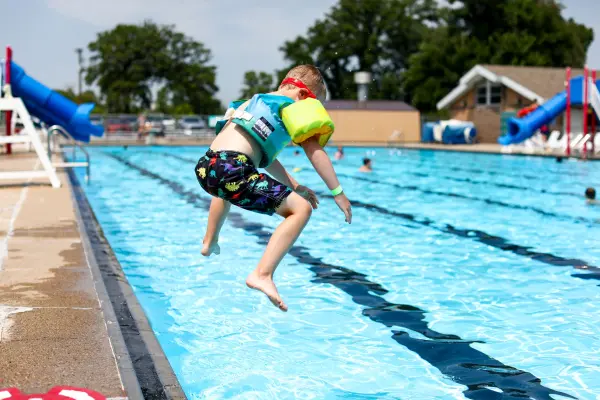 Aquatics fun: A child jumps into a blue swimming pool with water slides, wearing a life vest and dinosaur swim trunks.