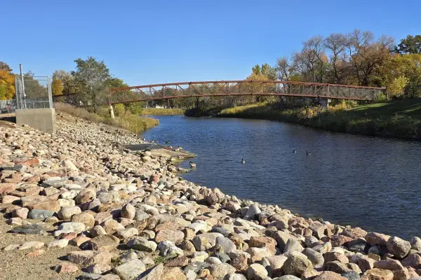 A metal pedestrian bridge crosses a calm river flanked by a rocky embankment and autumn trees under a clear blue sky.