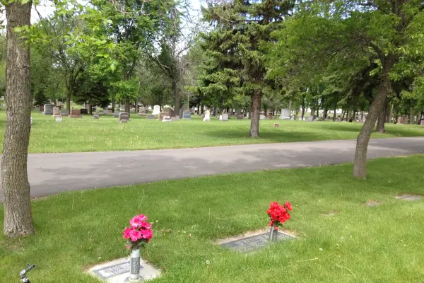 Paved pathway winding through a green cemetery with grave markers and vases of red and pink flowers.