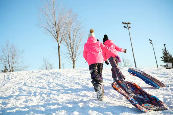Two children in bright pink coats walk up a snowy hill pulling plastic sleds on a sunny winter day.