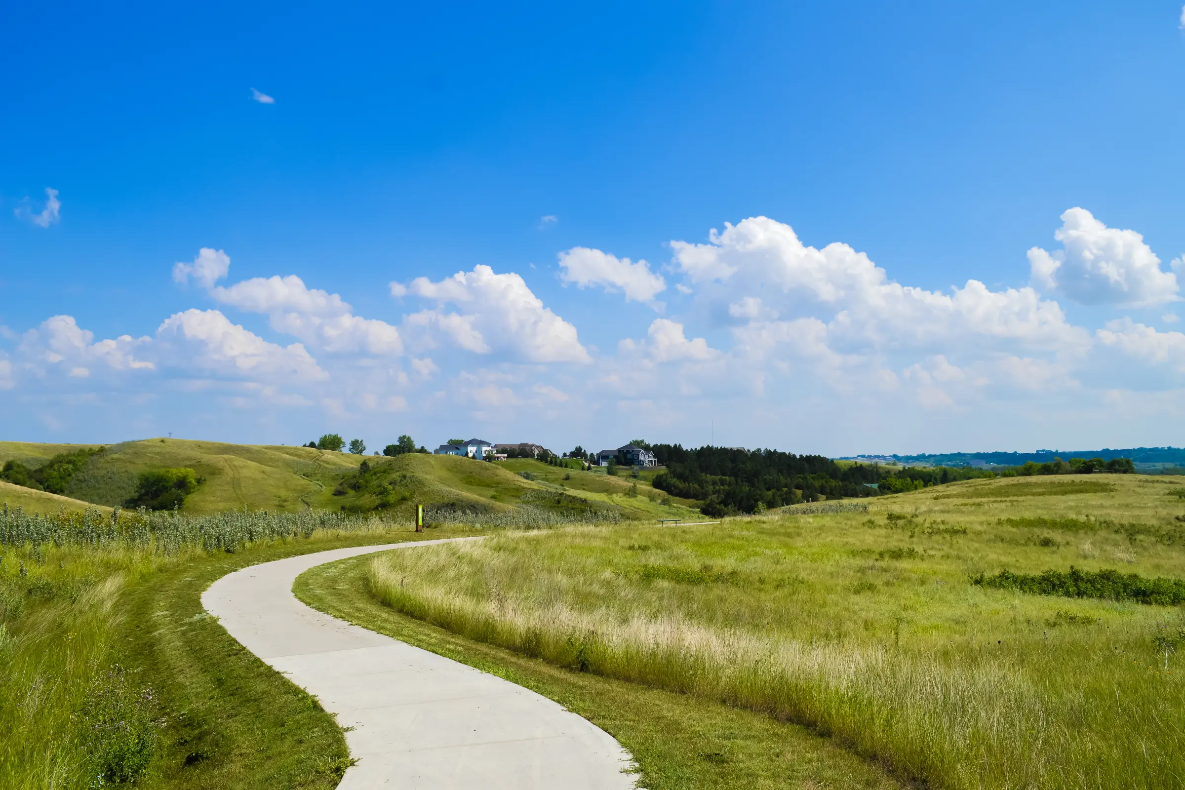 Paved trail curves through rolling green hills under a bright blue sky with scattered clouds.