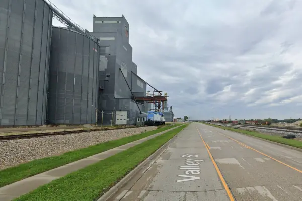 Industrial grain elevator with an adjacent train engine and paved sidewalk along Valley Street under a cloudy sky.