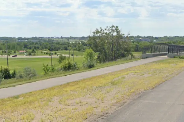 A paved walking/bike path along a highway with a trail-bridge and trees in the background under a blue sky with white clouds.