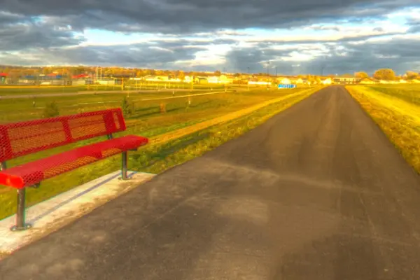 Screenshot from Google Maps of a stretch along the Souris Valley Golf Trail. Paved walking/biking path with a red metal bench nearby on a cloudy day.