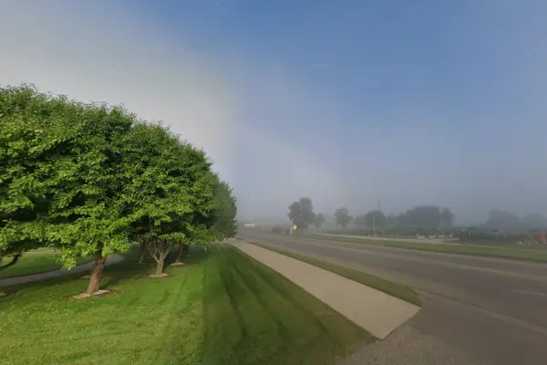 A broad paved sidewalk runs along an empty street lined with grass and large trees on one side.