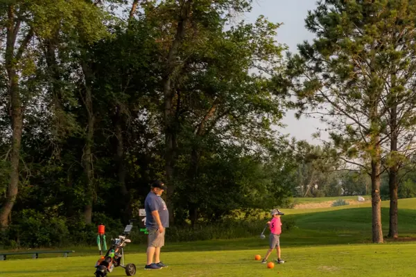 Man in a blue shirt watches a young child in a pink shirt swing a golf club on a sunny golf course.