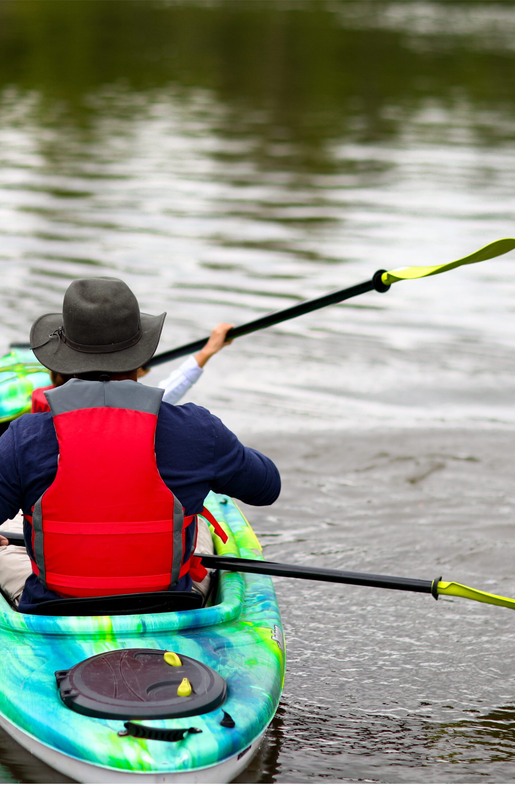 A person in a red life jacket paddles a green and blue kayak across calm water, creating gentle ripples.
