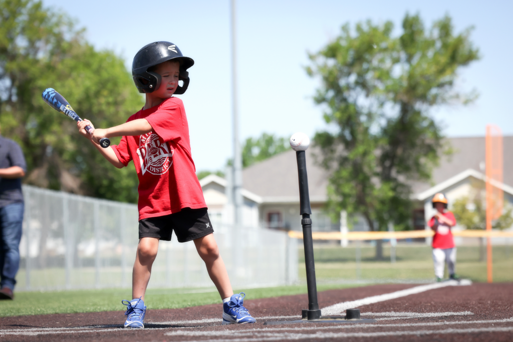Child in red shirt preparing to swing a baseball bat at a ball on a tee.