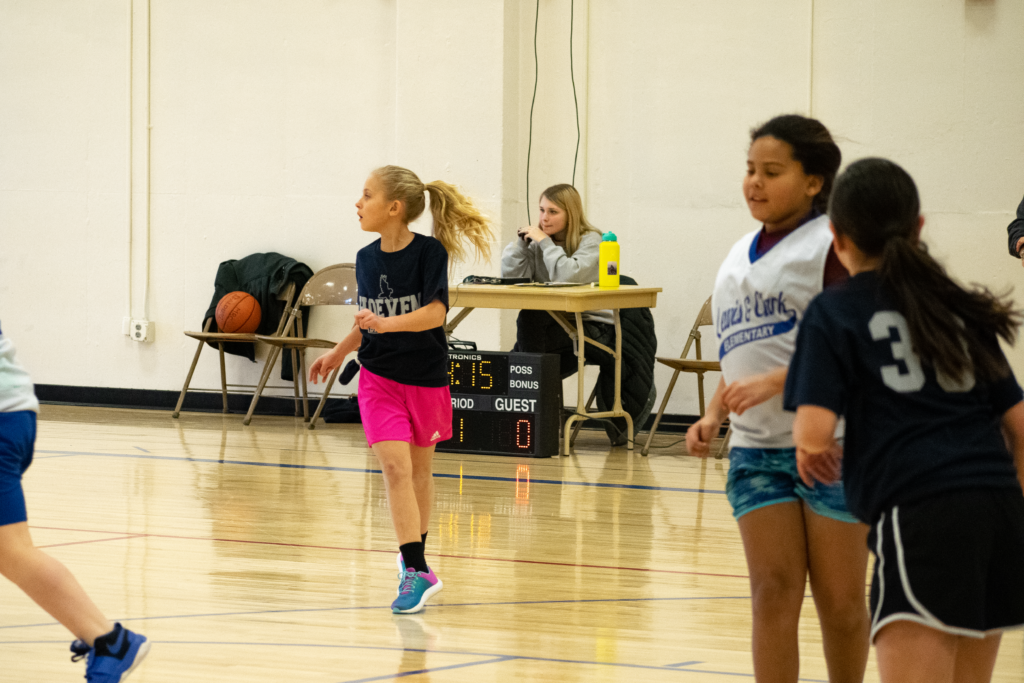 Young athletes in team jerseys move across a wooden basketball court near a scoring table and scoreboard.