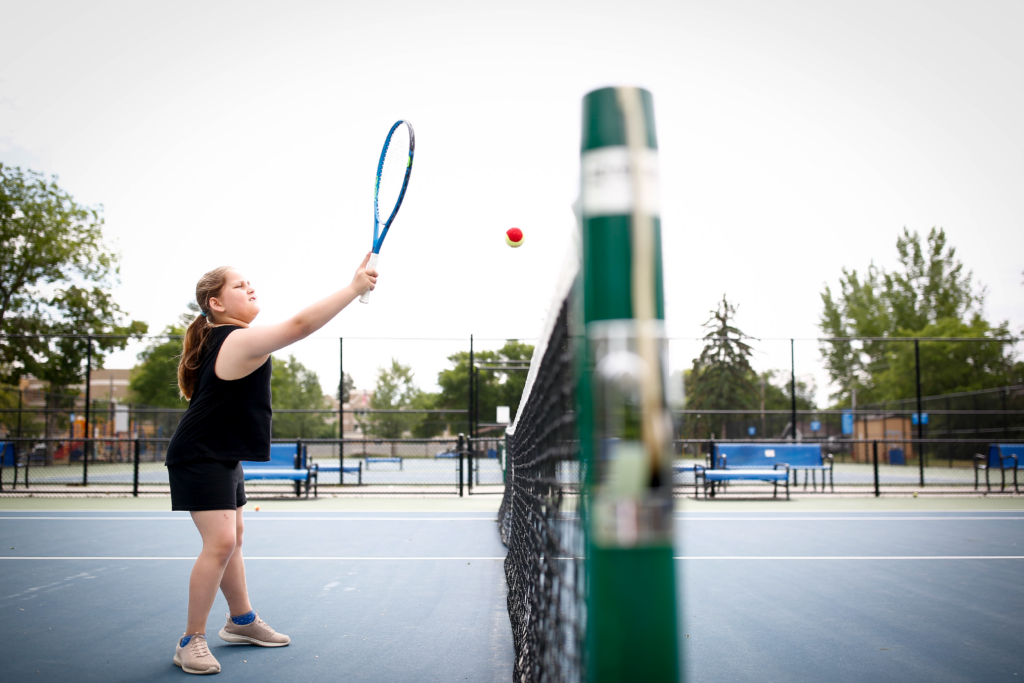 A player hits a red‑and‑yellow tennis ball near the net on an outdoor blue court.