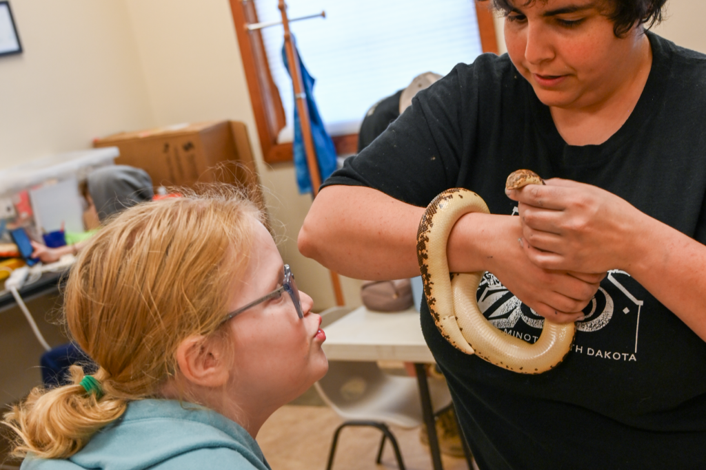Girl in glasses leans toward a light-colored snake coiled around an instructor's arm during a classroom presentation.
